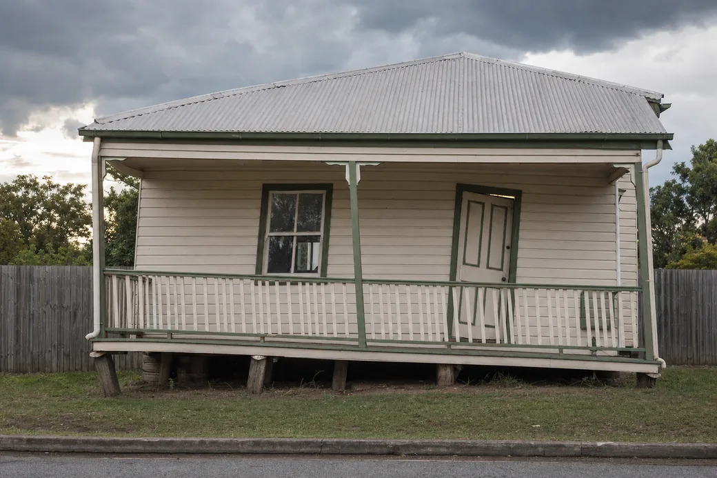 A low-set timber cottage whose verandah sags away from a straight timber fence behind it, with the front door and a window out of square — underpinning, Brisbane. Illustrative example.