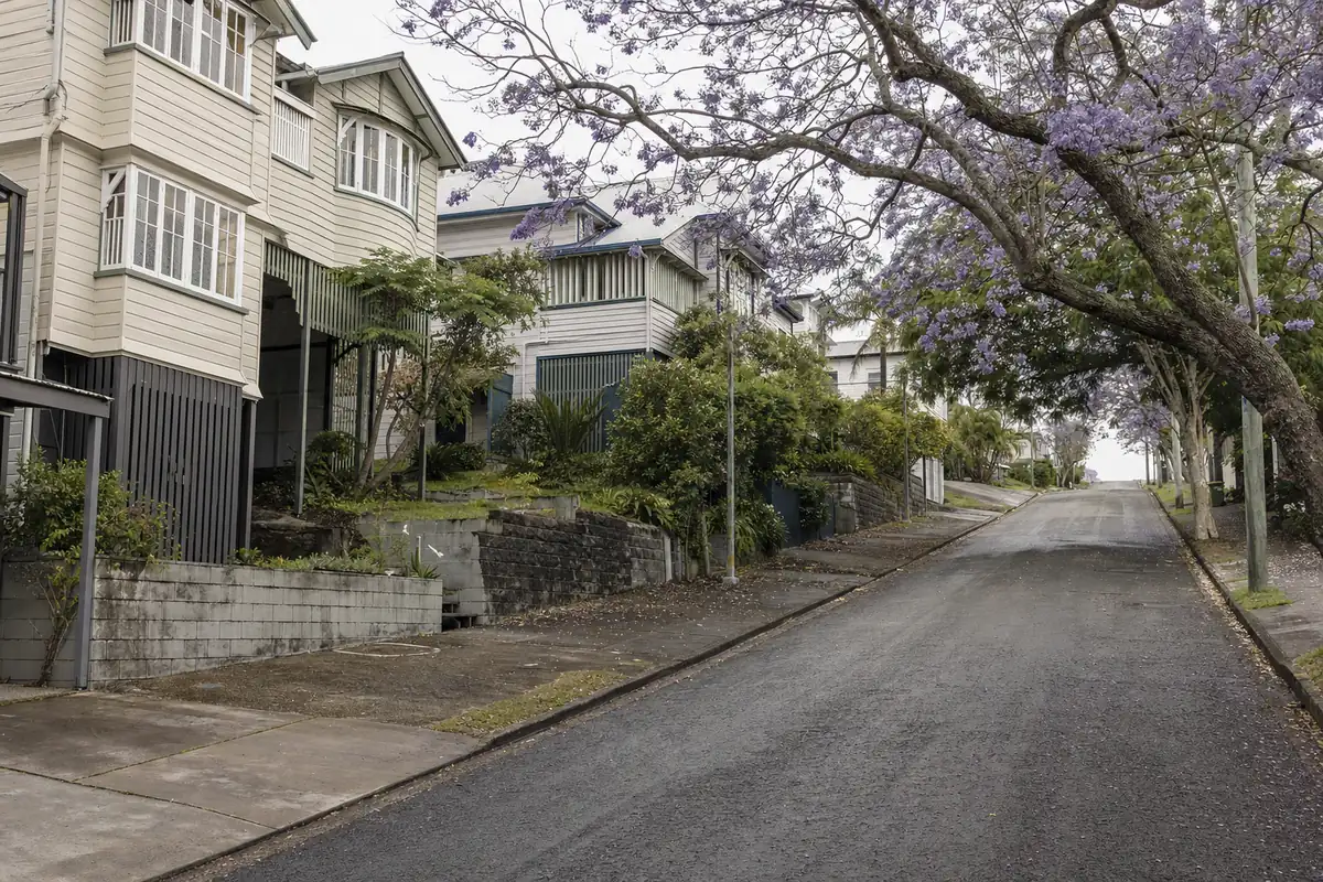 A steep street of high-set timber houses with open undercroft garages, low retaining walls at the footpath and a jacaranda in flower — underpinning, Brisbane. Illustrative example.