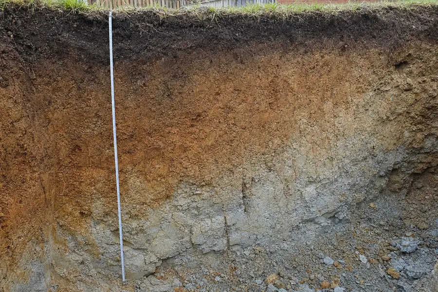 The cut face of an excavation with dark topsoil over orange-brown weathered material over pale grey broken rock, a tape measure laid against it — underpinning, Brisbane. Illustrative example.