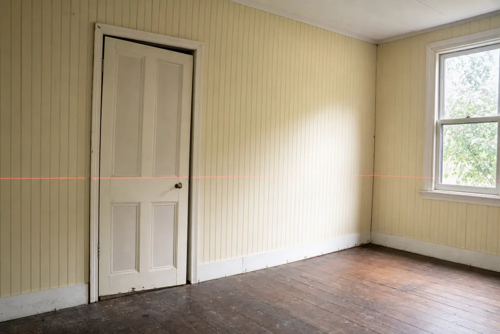 A red laser line running level across a timber-panelled wall past a panelled door that sits with a dark gap along its head — crack repair, Brisbane. Illustrative example.